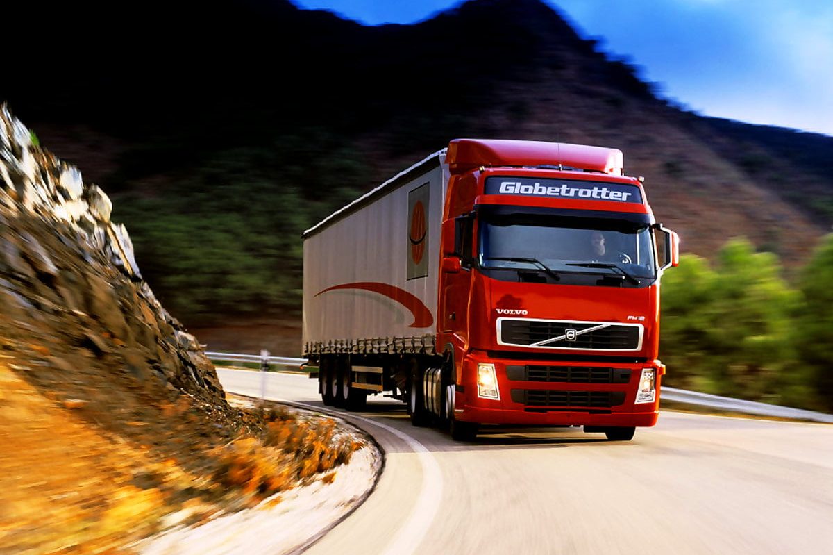 Körkortsintyg Röd lastbil kör på en kurvig väg omgiven av berg och grönska.